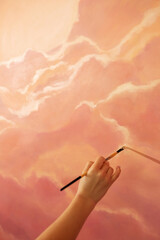 Using a peach fuzz color, the woman hand is painting with a brush a sunset or a sunrise clouds on the wall