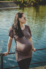 A pregnant girl stands on a bridge near a pond on a summer day