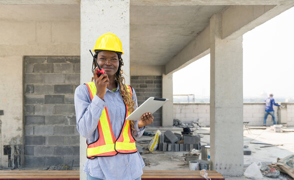 A builder in safety uniform standing at a building site, holding a walkie-talkie communicating with other team member. Tablet in her hand used for reviewing construction plan or checking schedule.