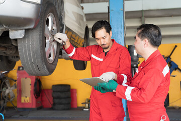 Two male mechanic working at garage. Asian male mechanics checking tire wheel underneath lifted car...