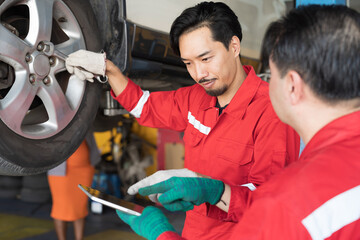 Two male mechanic working at garage. Asian male mechanics checking tire wheel underneath lifted car...