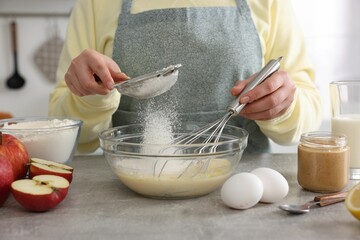 Woman whisking eggs while sieving flour at grey table indoors, closeup