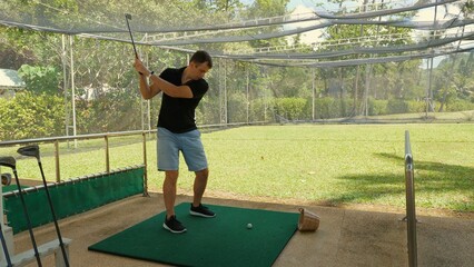 Focused man practicing golf swing on driving range with greenery in background. Outdoor recreational activities and sports training.