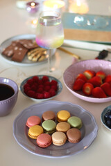Plate of pastel macarons, cookies and chocolate, cup of tea of coffee, glass of bubble water, various berries, books and accessories on the table. Selective focus, pastel colors.