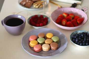 Plate of pastel macarons, cookies and chocolate, cup of tea of coffee, glass of bubble water, various berries, books and accessories on the table. Selective focus, pastel colors.