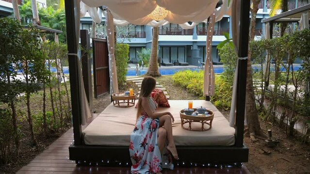 Woman enjoying serene moment alone on luxurious outdoor daybed surrounded by lush tropical greenery. Tranquility and peacefulness in exotic resort setting.