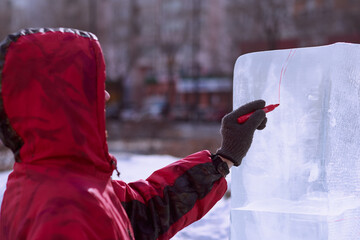 A sculptor draws a red line on an ice block. Preparing to create a sculpture at the city winter festival of artistic ice figures. - obrazy, fototapety, plakaty