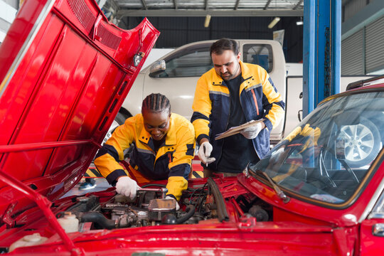 Two mechanic are working on a car with the hood open in an automotive shop or garage. They seem focused and engaged in diagnosing or repairing the vehicle's engine or other component.