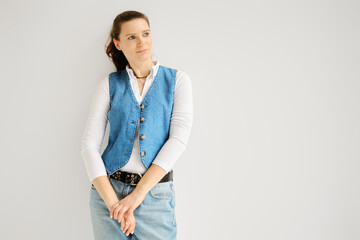 Relaxed woman in denim vest, jeans and cotton shirt leaning against white wall