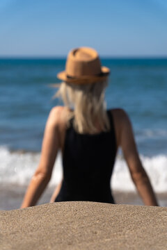 Close-up of an empty place on the sand on a blurred background of a young woman in a black swimsuit with a hat on the beach on a sunny day.Young girl is thinking and relaxing by the sea.Vertical photo