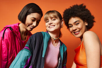 Three beautiful women of different ethnicities standing together in front of an orange backdrop.