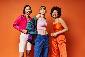 Three beautiful women of different ethnicities standing together, celebrating diversity on a vibrant orange studio background.