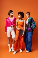 Three beautiful women of different ethnicities standing united in front of an orange background.