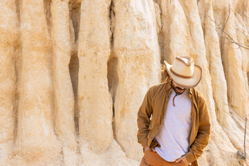A man wearing a cowboy hat stands in front of a large rock formation. He is wearing a brown jacket and a white shirt - Powered by Adobe