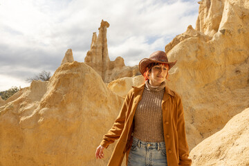 A woman in a brown jacket and hat stands in front of a large rock formation. Concept of adventure and exploration, as the woman is in a remote and rugged landscape - Powered by Adobe