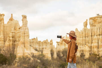A woman is taking a picture of a mountain range with a camera. She is wearing a brown jacket and a hat - Powered by Adobe