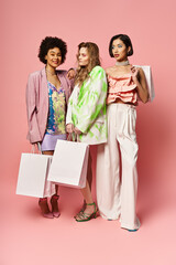 Three diverse women, Caucasian, Asian, and African American, standing together, holding shopping bags on a pink studio background.