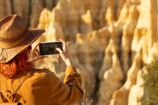 A woman wearing a straw hat is taking a picture of a mountain. The photo is in color and the woman is holding a cell phone