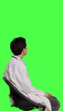 Profile Physician in white coat waiting for patients at consultations, feeling impatient sitting on a chair against greenscreen backdrop. Medic practitioner with stethoscope waits for people. Camera B