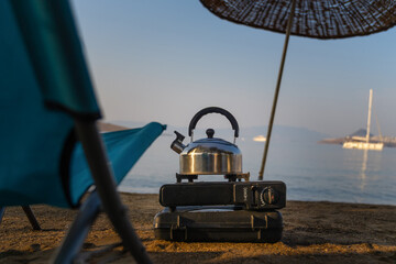 Metal kettle boiling on a portable gas stove against the background of a calm sea, a beach umbrella, a folding chair at sunrise. Travel and outdoor recreation.