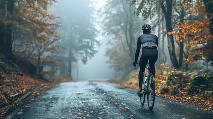 Foto op Plexiglas A cyclist in a black helmet and outfit riding a bicycle on a foggy wet road surrounded by autumn-colored trees. © iuricazac