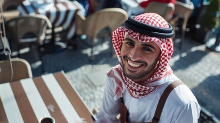 Smiling man in traditional Middle Eastern attire seated ata striped table surrounded by chairs and other patrons suggesting a social gathering or event. - obrazy, fototapety, plakaty