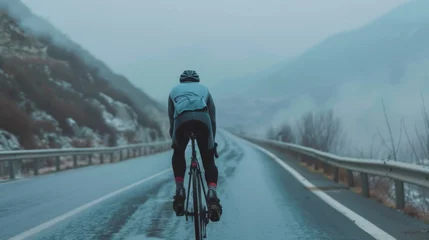 Fotobehang A cyclist in a blue jacket and black pants riding a bicycle on a wet road with foggy mountains in the background. © iuricazac