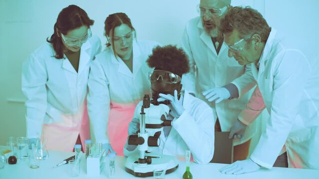 African university student analyzes a specimen under a microscope while surrounded by peers and professor in a lab