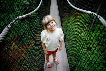 Cute little preschool girl walking on high tree-canopy trail with wooden walkway and ropeways on...