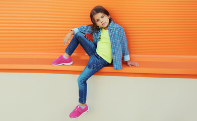 Stylish girl child in colorful clothes posing on city street on orange wall background