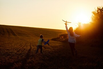 Running boy and girl holding two yellow and blue airplanes toy in the field during summer sunset. Kids dreams of flying and aviation.