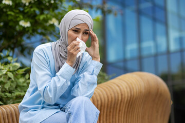 Sad muslim female sitting on wooden bench and covering face with paper tissue for blowing out. Sick...