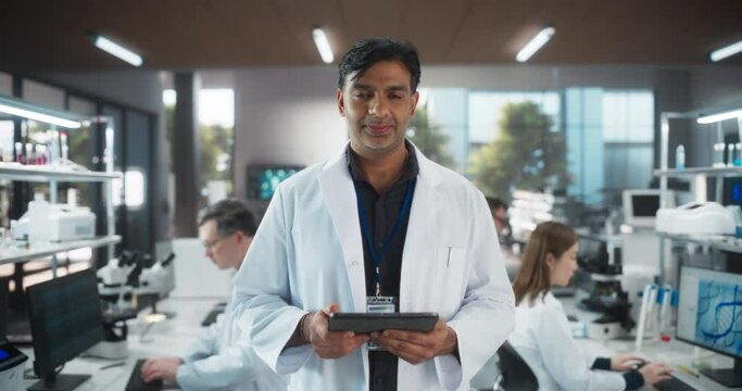 Portrait of an Indian Adult Male Scientist Using Tablet Computer, Looking at Camera and Smiling in a Laboratory. Professional Researcher Working as Bioengineer, Monitoring Technology Projects