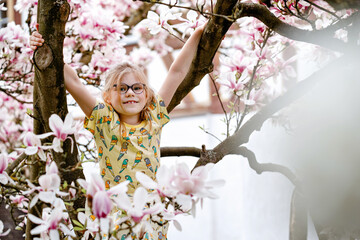Cute spring fashion preschool girl with glasses under blossom magnolia tree. Little happy child and...