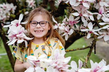 Cute spring fashion preschool girl with glasses under blossom magnolia tree. Little happy child and spring.