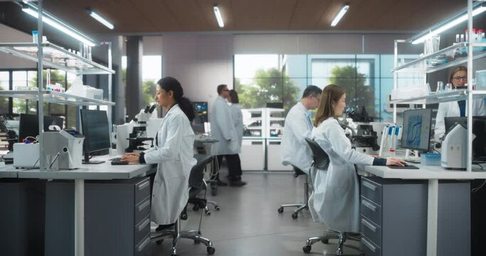 Team of Diverse Male and Female Medical Research Scientists Work Behind Desks on a New Generation Pharmaceutical Products in a Modern Laboratory. Colleagues Walking in the Background