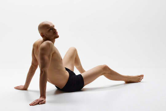 Shirtless man with perfect body curves posing with thoughtful expression in underwear on floor against grey studio background. Concept of men's health, self care, fashion and beauty, healthy lifestyle