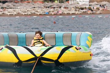 Young boy exudes joy while riding an inflatable tube towed by a boat in the ocean. Happy school child having fun in adventure water park on the sea.
