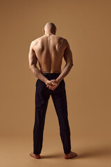 Rear view photo of bald man holding hands behind back and showing his muscular back against sandy studio background. Concept of men's health, self care, fashion and beauty, healthy lifestyle. Ad