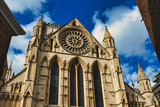 Gothic cathedral facade with rose window and spires against a blue sky with clouds, framed by trees in York, North Yorkshire, England.