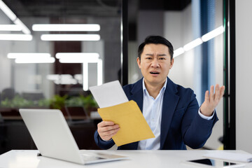 Confused asian man sitting at personal workspace and looking at camera while holding paper envelope with letter. Surprised manager spreading hands while receiving mail for unknown addressee. - obrazy, fototapety, plakaty
