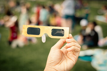 Hand holding paper solar eclipse with blurry crowd people watching totality show picnic yard, Dallas, Texas, April 8, scratch resistant polymer lenses filter out harmful ultraviolet, infrared ray
