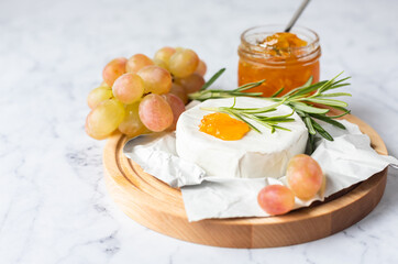 Delicious gourmet Camembert cheese garnished with orange jam, grapes, rosemary and basil on light background.