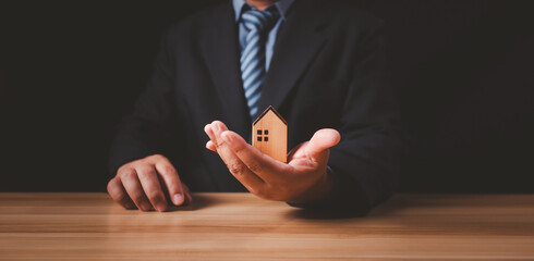 Businessman hand holding a wooden house on wooden table for housing and property concept, Insurance...