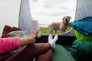 An unrecognizable woman in a tent drinks tea from a mug overlooking the lake. The dog is standing...