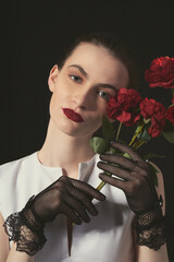 Close-up of a young woman in a white dress with bright makeup. The emphasis is on her hands in black translucent evening gloves decorated with lace. A girl is holding a bouquet of dark red roses in he