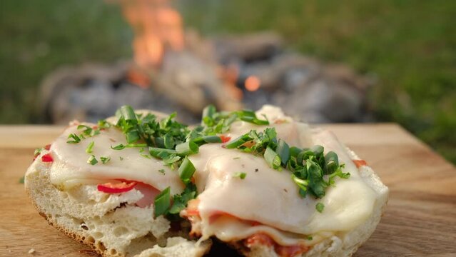 Close up of green parsley on hot baguette with sausage, onions and fresh tomato near campfire at warm spring or summer day. Process of cooking bread boat on wooden board at outdoor