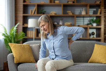 Mature lady feeling spinal muscles pain attack and holding back while squinting eyes on sofa with...