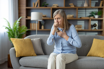 Senior stylish woman feeling sick and covering mouth with clenched fist for stopping cough while sitting on sofa with cushions. Unhealthy female feeling pain in chest and suffering from flu symptoms.