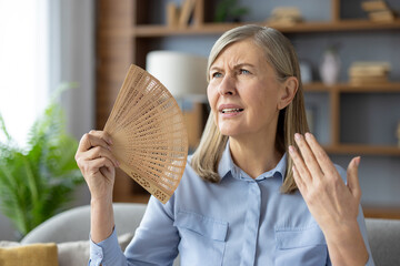 Disturbed woman waving with wooden hand fan to squinted face while sitting in overheated apartment....
